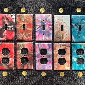 May include: A set of 12 decorative light switch and outlet covers featuring abstract, colorful, fluid art designs. The covers are labeled with letters A through N.