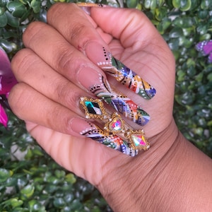 May include: Long, nude-coloured acrylic nails with intricate, colourful designs. The nails feature a mix of geometric shapes, swirls, and dots in shades of green, yellow, orange, blue, and black. Each nail is adorned with a gold-toned, jeweled accent.