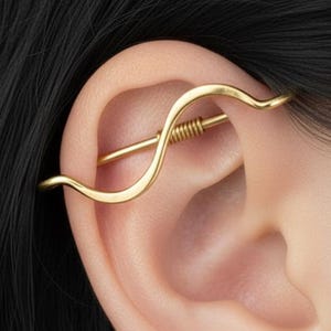 May include: A gold-toned industrial ear piercing jewelry piece featuring a smooth, wavy outer bar that curves across the upper ear cartilage, connected to a straight inner bar with a small, coiled spring-like detail in the center.