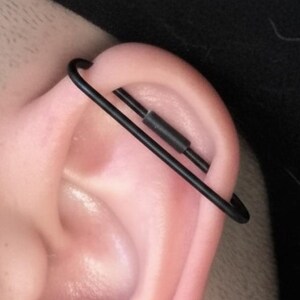 May include: A matte black industrial barbell earring featuring a long, thin metal bar that passes through two points of the upper ear cartilage, complemented by a large, smooth black ear gauge plug inserted into the earlobe below.