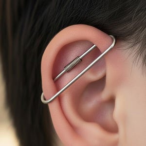 May include: A silver-toned industrial ear piercing consisting of a long, curved metal bar that spans the upper cartilage. The jewelry features a central coiled spring-like detail along the upper section of the bar, creating a distinct linear look.