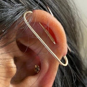 May include: A gold industrial barbell piercing with a long, thin, rectangular frame spanning the upper ear cartilage, paired with a small gold flower stud in the conch and three gold hoop earrings of varying sizes along the earlobe.