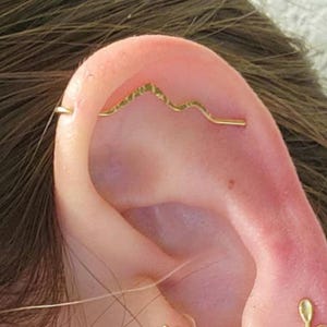 May include: A gold-toned ear jewelry set featuring a mountain range industrial bar, a sun stud with a small red center, a leaf-shaped stud, and a simple hoop earring. These pieces are arranged along the cartilage and lobe of the ear.