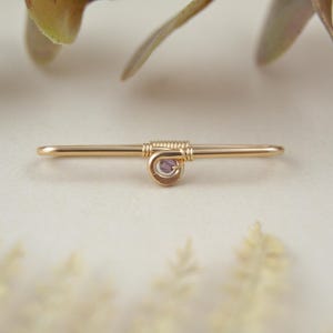 May include: A gold-toned metal industrial barbell piercing jewelry featuring a straight bar with a central spiral wire-wrapped design. A small, round, light purple gemstone is set within the center of the wire coil, creating a delicate accent.