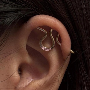 Puede incluir: Un pendiente tipo ear cuff de tono dorado con un diseño ondulado continuo que se envuelve alrededor del cartílago superior de la oreja. El metal tiene una superficie pulida y reflectante que se curva en una forma de bucle redondeado.