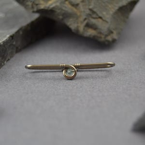 May include: A horizontal metal bar brooch featuring a central coiled wire design that secures a small, round, pale blue gemstone. The metal has a muted, dark bronze-toned finish with visible wire wrapping details at the center point of the bar.