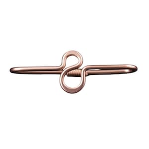 May include: A copper-colored metal hair barrette with a unique, abstract design. The barrette features a central looped element and straight, elongated ends. The metal has a polished finish, reflecting light.