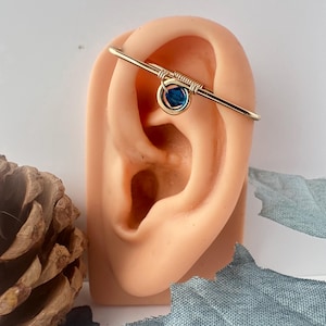 May include: A gold-toned industrial barbell ear piercing featuring a central circular charm wrapped in wire. A small, faceted deep blue crystal bead is suspended within the center of the gold ring, creating a distinct contrast against the metallic gold wire.