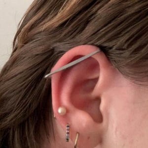 May include: A silver-toned industrial barbell piercing bar extending across the upper ear cartilage. Below this, a single white spherical stud earring is positioned, followed by a small silver hoop earring and a thin gold-toned hoop earring.