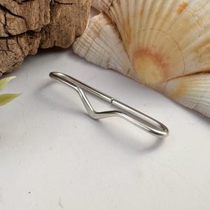 May include: A silver-toned metal tie bar featuring a smooth, elongated, and slightly wavy design. The accessory has a polished finish and a simple, minimalist construction with a single continuous loop that forms the clip mechanism.