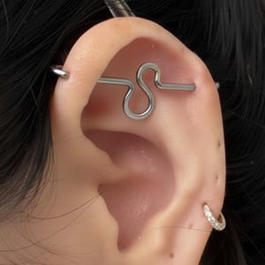 May include: A silver-toned industrial barbell piercing jewelry featuring a central S-shaped curve, positioned horizontally across the upper ear cartilage, accompanied by a small silver hoop earring with clear stones in the earlobe.