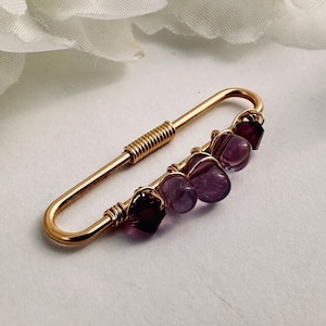 May include: A gold-toned metal safety pin brooch featuring a series of five beads attached with wire wrapping. The beads include two dark purple faceted crystals at the ends and three smooth, round, translucent light purple amethyst stones.
