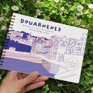 Peut inclure: Un carnet de coloriage à spirales avec le titre "DOUARNENEZ" en bleu. La couverture présente une illustration d'une ville côtière aux tons violets et roses. Le texte "Balade sur le port du Rosmeur" et "Carnet de coloriages sensibles" est également visible.