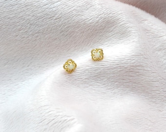 Puede incluir: Un par de pendientes de botón dorados. Cada pendiente presenta un diseño de trébol de cuatro hojas con un pequeño centro de piedra preciosa blanca. Los pendientes están colocados sobre un fondo blanco suave.