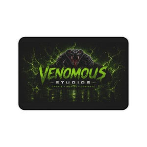 May include: A rectangular black desk mat featuring a graphic of a black snake with red eyes centred above the lime green text VENOMOUS STUDIOS. Below the main text, the words CREATE, INSPIRE, DOMINATE appear in a smaller, white font.