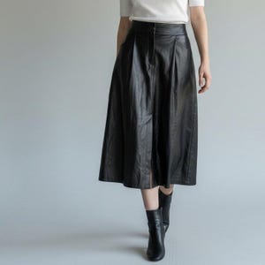 May include: A black midi-length skirt with a high waist, featuring a smooth finish and subtle vertical pleats. The skirt has a structured waistband with a central button closure and falls in a flared silhouette, paired with black ankle boots.