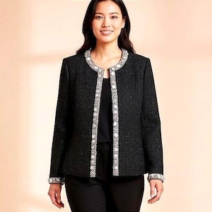 Könnte beinhalten: Schwarze Jacke mit strukturiertem Gewebe, rundem Ausschnitt und vorderer Zierleiste mit weißen und silbernen Akzenten. Die Jacke hat lange Ärmel mit passenden verzierten Manschetten. Die Frau trägt eine schwarze Hose.