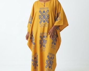 Pode incluir: Um vestido caftan longo até ao chão em amarelo mostarda com decote em gota e mangas largas. A peça é decorada com um padrão repetitivo de formas geométricas azul-escuras e motivos lineares dispostos em colunas verticais no tecido.
