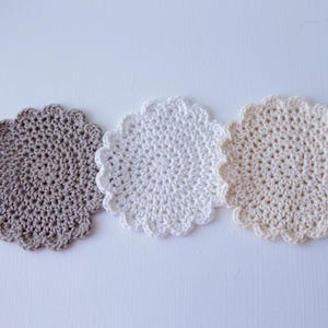 Op de afbeelding: Drie gehaakte onderzetters in verschillende tinten bruin, beige en wit. De onderzetters zijn rond en hebben een gegolfde rand.