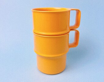 May include: Two stacked orange ceramic mugs with handles. The mugs are a bright, solid orange color.