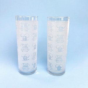 May include: Two clear glass drinking glasses with a white design. The design features a cartoon mouse holding a donut and the text "The Little Donuts" and "The Little Beignets".