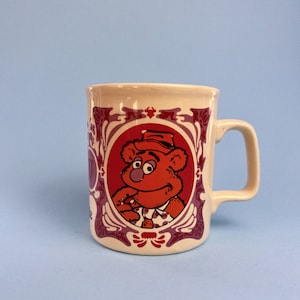 May include: A white ceramic mug with a red and purple design featuring a cartoon bear wearing a hat and a shirt. The bear is in a circular frame with a floral design.