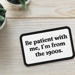 May include: Two small potted succulents and a white rectangle with black border. The rectangle has the text: "Be patient with me, I'm from the 1900s." The plants are green and the background is light wood.