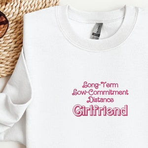 May include: White sweatshirt with the text "Long-Term Low-Commitment Distance Girlfriend" in pink.
