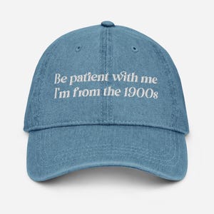 May include: A light blue denim baseball cap with the text "Be patient with me I'm from the 1900s" embroidered in white. The cap has a curved brim and an adjustable strap.