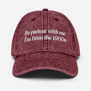 May include: A maroon baseball cap with a curved brim. The cap features the embroidered text "Be patient with me I'm from the 1900s" in white. The cap has a vintage, worn look.
