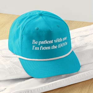 May include: A turquoise baseball cap with a white rope detail along the brim. The cap features the embroidered text, "Be patient with me I'm from the 1900s" in white. The cap is resting on a white surface.
