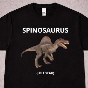 Puede incluir: Camiseta negra con una ilustración de un dinosaurio Spinosaurus. La palabra "SPINOSAURUS" está impresa encima del dinosaurio, con "(HELL YEAH)" debajo. El dinosaurio está representado en tonos marrones y grises.