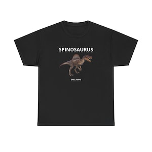May include: Black t-shirt featuring a Spinosaurus dinosaur illustration. The text "SPINOSAURUS" is above the dinosaur, with "(HELL YEAH)" below. The dinosaur is depicted in shades of brown and gray, with an open mouth.