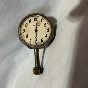 May include: An antique Tacoma clock with a white face, black numerals, and a metal casing. The clock has a stem and a textured knob. The hands point to 6 o'clock. The clock is likely vintage.