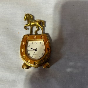 May include: Gold-toned clock shaped like a horseshoe with a gold horse figurine on top. The clock face is white with black hands and black numbers.
