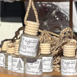 May include: A collection of small glass car diffuser bottles with light wood caps and jute hanging cords. Each bottle features a white label with black text reading car diffuser, candlelit tree, and Hursh Homegrown, plus a stocking icon.
