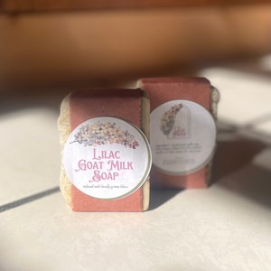 May include: Two bars of Lilac Goat Milk Soap with a light beige and mauve color scheme. Each bar is wrapped with a mauve paper band and a round label featuring lilac flowers and the text "Lilac Goat Milk Soap".