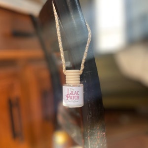 May include: A small, clear glass car diffuser with a wooden cap and a woven cord. The label reads "The Lilac Patch Car Diffuser" in a pink script font. The diffuser is suspended against a blurred background.