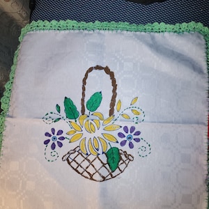 May include: A white crocheted square with a green border featuring a hand-painted design of a brown basket filled with yellow flowers and green leaves. The flowers have purple centers.