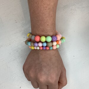 May include: A multi-coloured beaded bracelet with pastel shades of pink, blue, green, yellow, and brown. The bracelet is made of round beads and is stacked on a person's wrist.