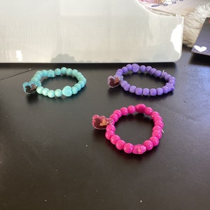 May include: Three beaded bracelets with heart-shaped charms. The bracelets are turquoise, purple, and pink.
