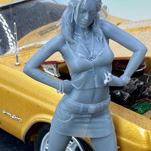 May include: A gray plastic figurine of a woman wearing a short skirt and a tank top, standing next to a yellow car. The figurine is posed with one hand on her hip and the other holding a wrench.