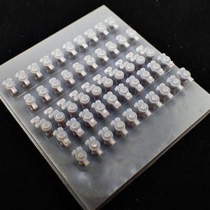 May include: A sheet of small, grey plastic components, possibly for model building or crafting. Each piece has a circular top and a stem, arranged in neat rows and columns on a rectangular base. The components are uniform in size and shape.