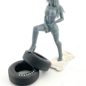 Könnte beinhalten: Graue Figur einer Frau in bauchfreiem Top, Shorts und Stiefeln, die einen Fuß auf einem schwarzen Reifen posiert. Ein weiterer Reifen befindet sich daneben. Die Reifen tragen das Wort "GOODYEAR".