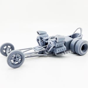 Puede incluir: Modelo gris de dragster con dos motores grandes, una barra antivuelco y neumáticos traseros grandes. Las ruedas delanteras son más pequeñas y tienen un diseño clásico de radios. El vehículo está sobre un fondo blanco.
