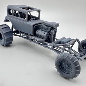 May include: A grey model of a custom hot rod with large, knobbly tyres. The vehicle has a detailed engine, roll cage, and a skeletal frame. The car is designed for off-road use and features a unique, open-top design.