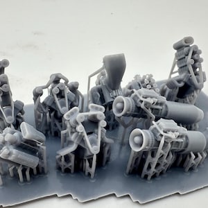 May include: A selection of small, grey, 3D-printed model engine components. The parts include various engine blocks, exhaust systems, and other mechanical elements, all arranged on a grey base.