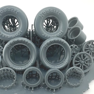 May include: A selection of grey miniature racing tyres and wheels, including several sets of tyres with the word "Hoosier" printed on the sidewalls. The wheels have intricate spoke designs, and the parts are arranged on a grey base.