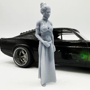 Puede incluir: Una figurita de plástico gris de una mujer con un vestido blanco y un collar plateado. La figurita está de pie junto a un coche negro con detalles verdes.