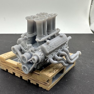 Op de afbeelding: Een grijs 3D-geprint model van een V8-motor op een houten pallet. De motor heeft vier inlaatspruitstukken en de tekst "5.72" is zichtbaar aan de zijkant.